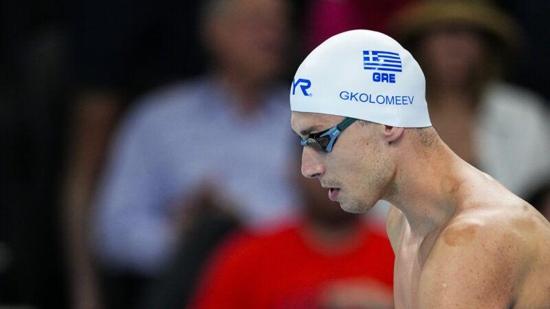 A man in the foreground, wearing swimming goggles and a swimming cap with the Greek flag on it. A few people are in the audience in the background, sitting. | Oscar J. Barroso/ZUMAPRESS/Newscom