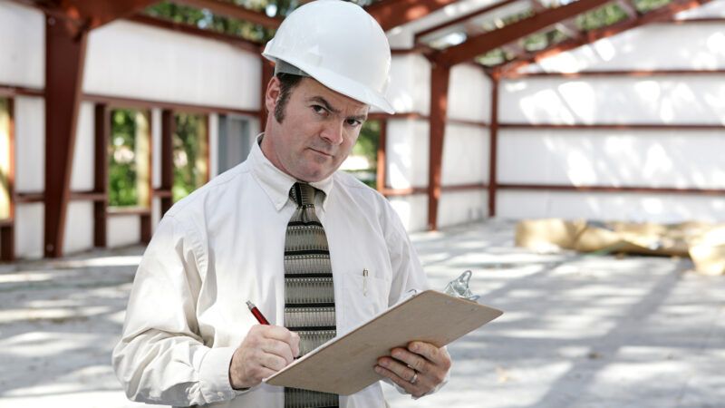 A code inspector in a hard hat stands in a building under construction, holding a clipboard and giving the camera a suspicous look. | Brian T. Young | Dreamstime.com