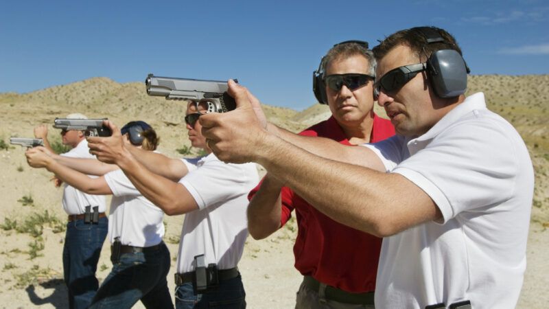 A line of people holds pistols ready to fire, with ear protection. An instructor coaches one of them. | Photographerlondon | Dreamstime.com