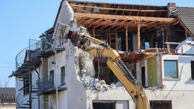 The arm of an excavator demolishes a residential home by tearing into the side, exposing an upper floor. | Hanna Kaplia | Dreamstime.com