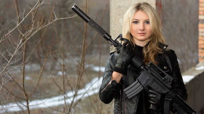 A blonde girl in tactical gear holds a military-style rifle, with a suppressor attached to the end of the barrel, across her torso. | Sergey Sukhorukov | Dreamstime.com