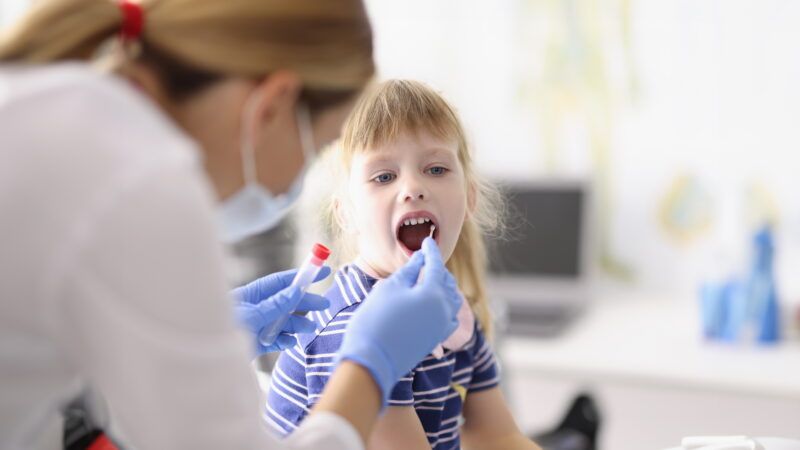 A female laboratory technician in protective gear swabs inside a little girl's cheek for a biological sample. | Kuprevich | Dreamstime.com