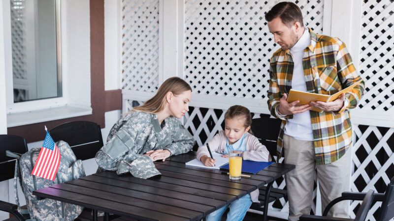 A young girl does homework at an outdoor table, surrounded by her mother—who is wearing combat fatigues—and her father. | Lightfieldstudiosprod | Dreamstime.com