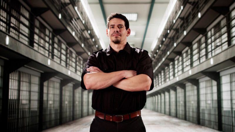 A prison guard in uniform stands with his arms folded, between rows of cells. | Andrey Popov | Dreamstime.com