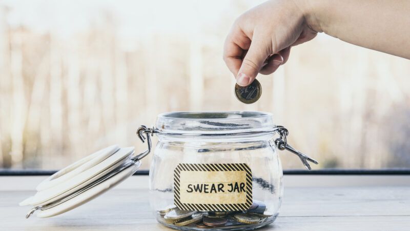 A child's hand places a coin into a clear jar filled with money, labeled "Swear Jar." | Helinloik | Dreamstime.com
