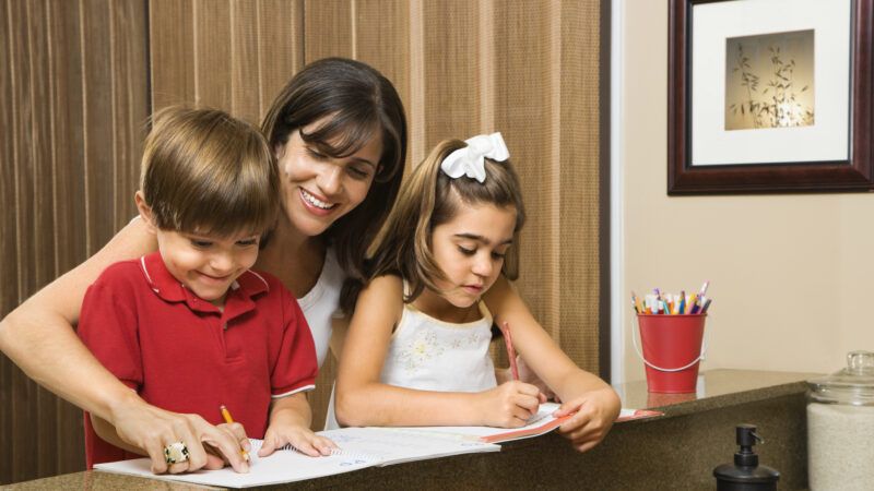 Two kids write on papers at a table while a smiling woman looks over their shoulder | ID 4246326 © Iofoto | Dreamstime.com