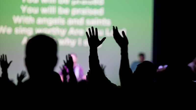Hands raised during a religious service with a band and lyrics on a big screen in the background | https://www.dreamstime.com/christians-worshipping-church-christians-raising-their-hands-church-worshipping-jesus-their-religious-service-image108407356