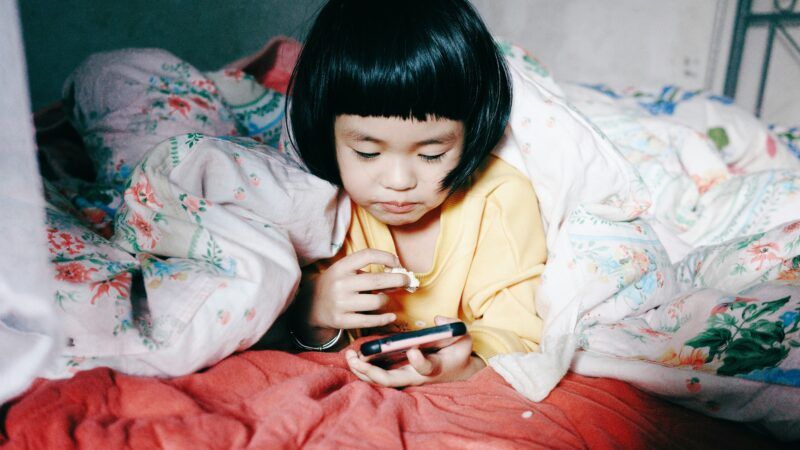 A young child using a smartphone | Photo by <a href="https://unsplash.com/@pandales?utm_content=creditCopyText&utm_medium=referral&utm_source=unsplash">Frank Ching</a> on <a href="https://unsplash.com/photos/a-little-girl-laying-in-bed-looking-at-a-cell-phone-KxN1IBdtxH0?utm_content=creditCopyText&utm_medium=referral&utm_source=unsplash">Unsplash</a>       
