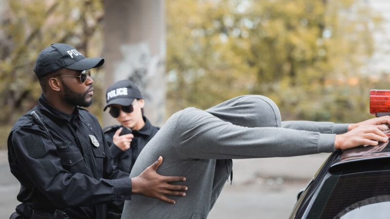 A police officer frisks a suspect in a grey hoodie, who has his hands on top of his car. | Lightfieldstudiosprod | Dreamstime.com
