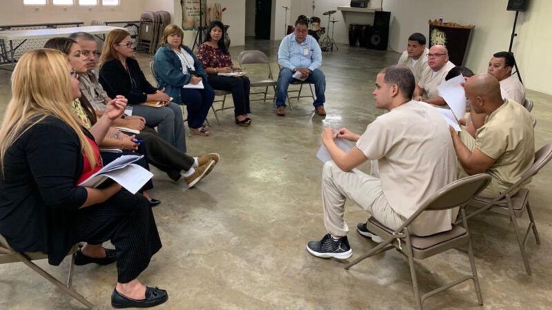 A group of people sitting in a circle of folding chairs; some of them are wearing inmate uniforms A group of people sitting in a circle of folding chairs; some of them are wearing inmate uniforms | Photo: Comisión de Desarrollo Cooperativo