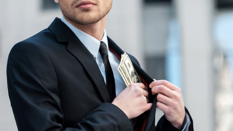 A man in a business suit stuffs cash into his jacket pocket. | Gstockstudio1 | Dreamstime.com