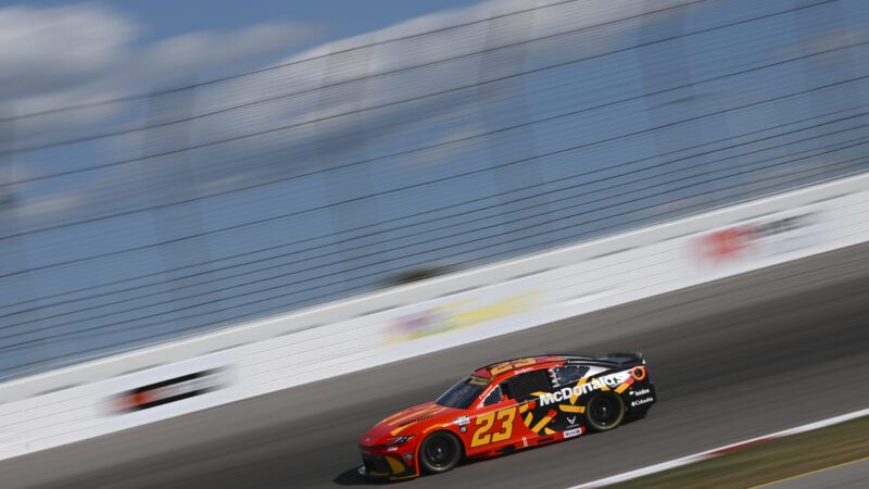 Side profile shot of a NASCAR stock car with 23 on the side and the word McDonald's and the fast food company's golden arches on the hood, while the car is traveling at high speed in front of a white wall and a partly cloudy blue sky. | Michael Allio/Icon Sportswire 116/Michael Allio/Icon Sportswire/Newscom