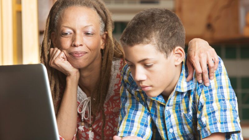 A mother puts her arm around her son's shoulder as he works on a laptop at the kitchen table. | Creatista | Dreamstime.com