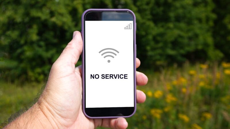 A man's hand holds a cell phone in a field. The phone's screen says "No Service," with a grey WiFi symbol. | Maskarad | Dreamstime.com
