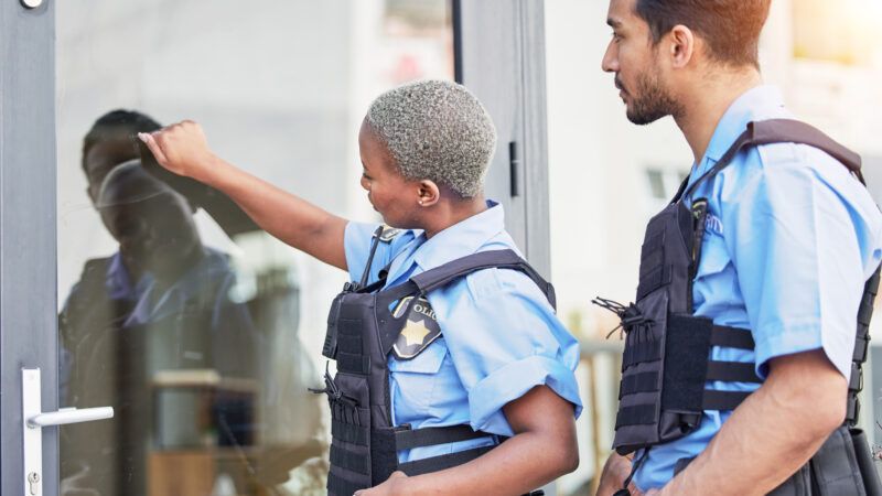 A black female police officer knocks on a residential storm door while a white male officer stands behind her. They both wear uniforms and body armor. | Yuri  Arcurs | Dreamstime.com