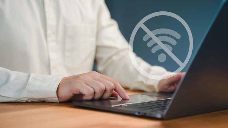 A man uses a laptop. A floating WiFi symbol with a circle and a line through it signifies an inability to connect to the internet. | Parin Kiratiatthakun | Dreamstime.com