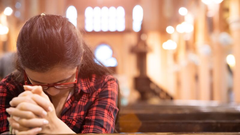 A young woman in church clasps her hands and bows her head in prayer. | Siam Pukkato | Dreamstime.com