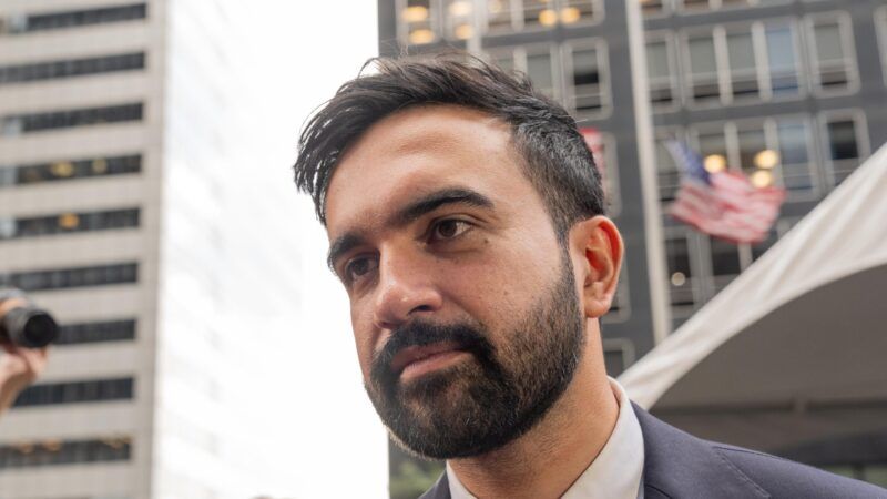 Zohran Mamdani photographed from below diagonal angle, with tall buildings behind him and a small American flag waving in the wind. Zohran Mamdani photographed from below diagonal angle, with tall buildings behind him and a small American flag waving in the wind. | Lev Radin/ZUMAPRESS/Newscom