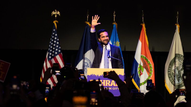 Zohran Mamdani speaks to a crowd behind a podium with his campaign sign attached and with flags in the background | Lev Radin/Sipa USA/Newscom