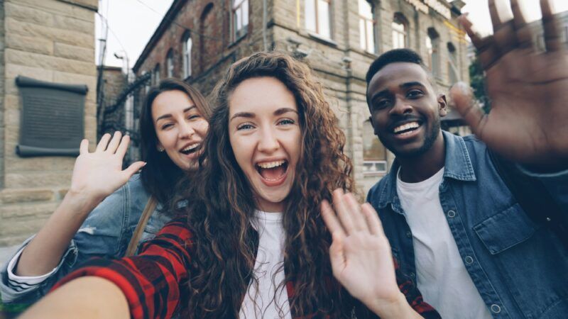 Young adults smiling and waving in a selfie | Photo by <a href="https://unsplash.com/@silverkblack?utm_source=unsplash&utm_medium=referral&utm_content=creditCopyText">Vitaly Gariev</a> on <a href="https://unsplash.com/photos/friends-wave-excitedly-taking-a-selfie-together-rtLU82A-K6w?utm_source=unsplash&utm_medium=referral&utm_content=creditCopyText">Unsplash</a>       