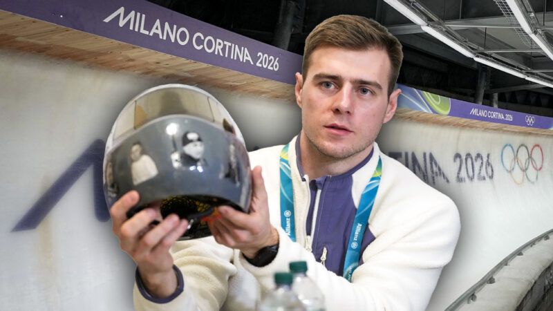 In the background is the curve of a sliding track with the Milano Cortina 2026 Olympics branding, in front of the background is a separate image of Vladyslav Heraskevych holding up a gray helmet with several faces on it. | Illustration: Kyodonews/ZUMAPRESS/Imagine China/Newscom