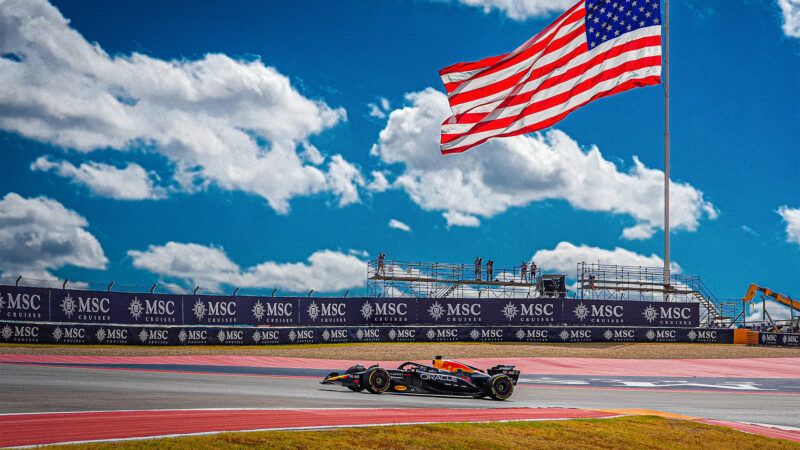 In front of a background of a blue sky and scattered white clouds, a Red Bull-branded Formula 1 car turns a corner beneath a massive American flag. | Credit: Alessio De Marco/Sipa USA/Newscom