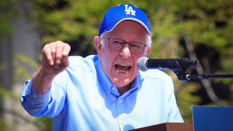 Sen. Bernie Sanders delivering a speech into a microphone, wearing a Los Angeles Dodgers hat. There is a background of trees behind him. | Photo: Yolanda Ruiz/ZUMAPRESS/Newscom