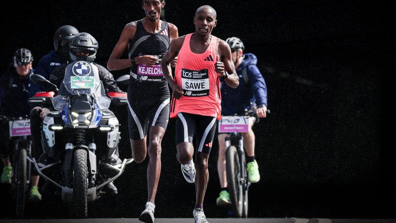 In front of a black background, Sabastian Sawe and Yomif Kejelcha run toward the camera as two bicycles and two motorcycles trail behind them. | Credit: Henning von Jagow / Action Plus/Newscom