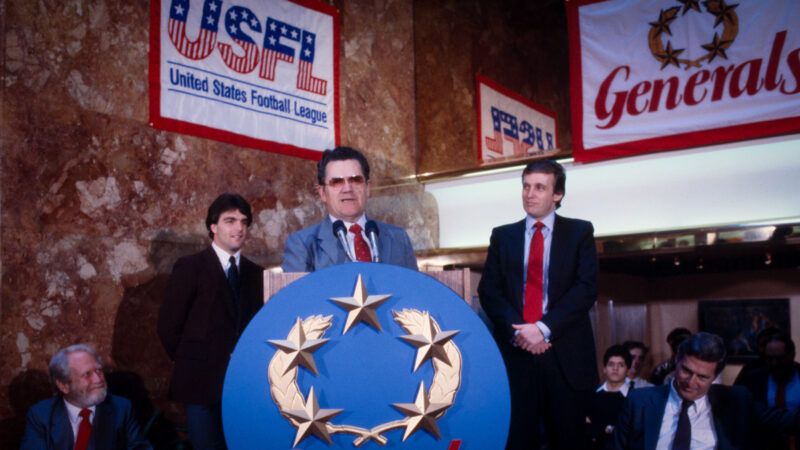 New Jersey Generals coach speaks at a lectern during a press conference, flanked by team owner Donald Trump and quarterback Doug Flutie. | Photo: Bernard Gotfryd/Wikimedia Commons