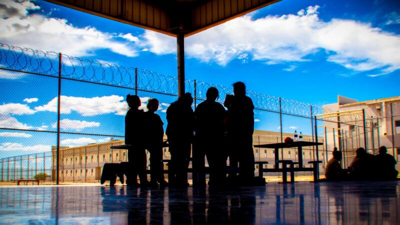 A group of people huddled in the shade beneath a roof, standing next to a barbed wire fence. | Photo: U.S. Immigration and Customs Enforcement