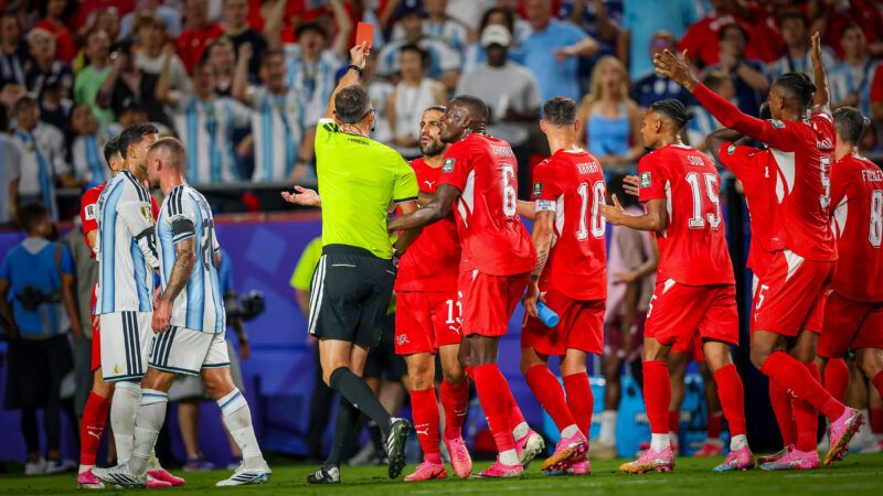 On a soccer field, a referee holds up a red card. A group of players in red jerseys is to the referees right, and two players in white and light blue jerseys are on his left. | Roman Puno/Cal Sport Media/Newscom