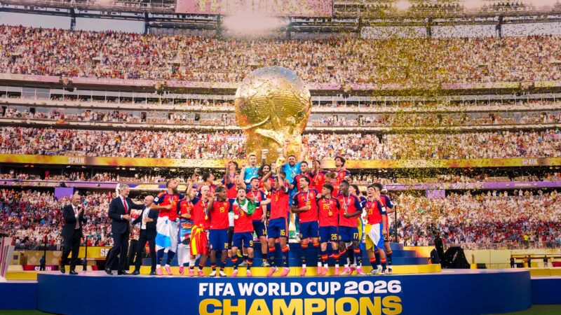 The Spanish soccer team celebrates on a stage in front of a stadium crowd and a large, golden World Cup trophy in the background. | Photo: EyePress/Newscom