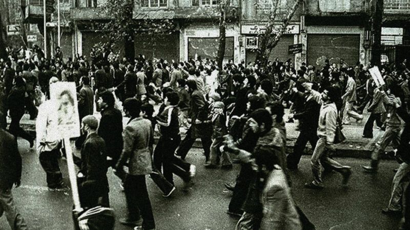 A black-and-white photo of people marching in a street, all toward the left side of the photo frame. A few of them hold signs on posts. | Wikimedia Commons