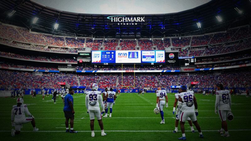 A group of players in football jerseys stands on the field, spread out, all facing a far-off end zone. The stands rise above them with speckles of red, white, and blue, with a video scoreboard and a logo that says "HIGHMARK STADIUM." | Bjorn Franke/Buffalo Bills