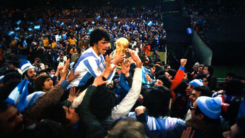An Argentine soccer player holding the World Cup trophy and being held up by teammates and fans | Mirror Syndication International/Mirrorpix via Getty