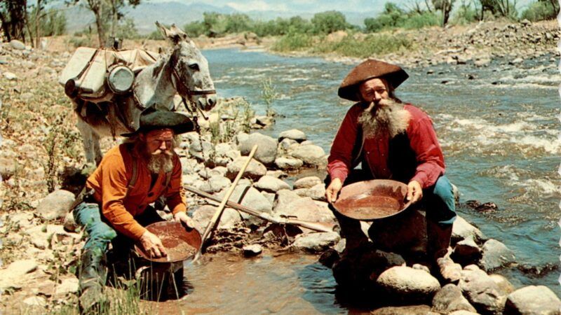A photo of two men holding gold pans with a donkey beside a rocky stream | Published by Royal Pictures, colored by Merle Porter; Public domain