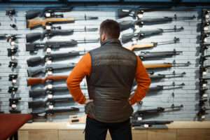 A man in an orange shirt and a vest stands with his back to the camera in front of a wall of long guns, for sale and on display. | Nomadsoul1 | Dreamstime.com