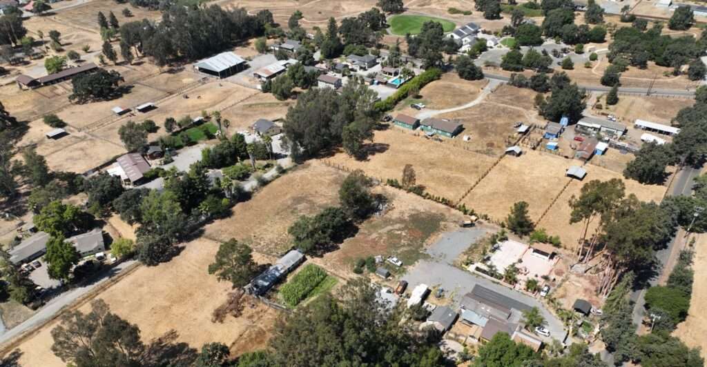 From above, a smattering of homes and other buildings surrounded by brown/yellow grass and some green trees.