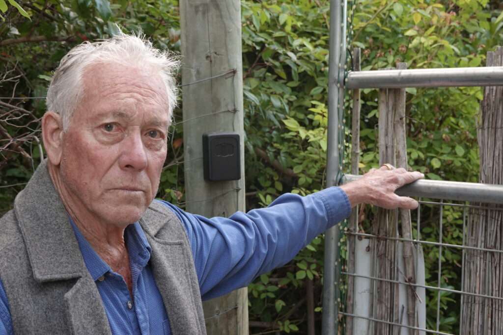 An older man with white hair puts his left hand on top of a tall gate/fence.