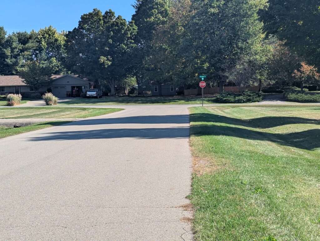 A residential street with shadows from two large trees over a lawn and the road, with a STOP sign further off in the distance.
