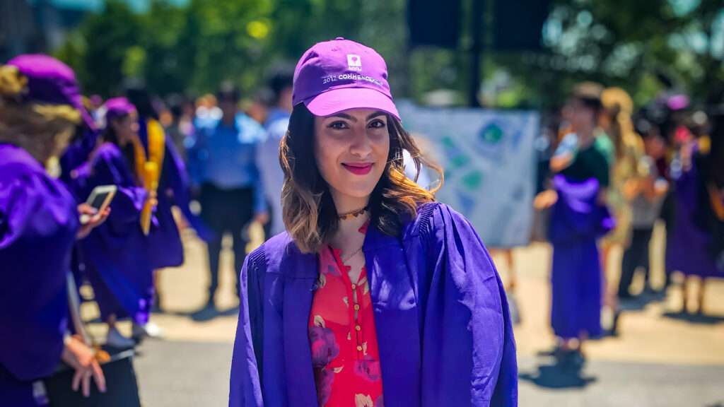 A woman wearing purple graduation robes and a purple baseball hat smiles, with a smattering of other people in purple graduation robes standing in the area.