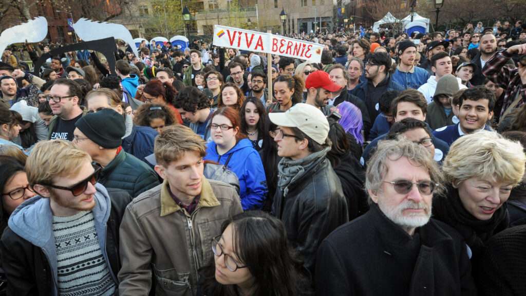 Many people are gathered in cold weather clothes in a wide open area surrounded by tall buildings—one of them is holding up a sign that says "ViVA BERNIE" with a Venezuelan flag on it.