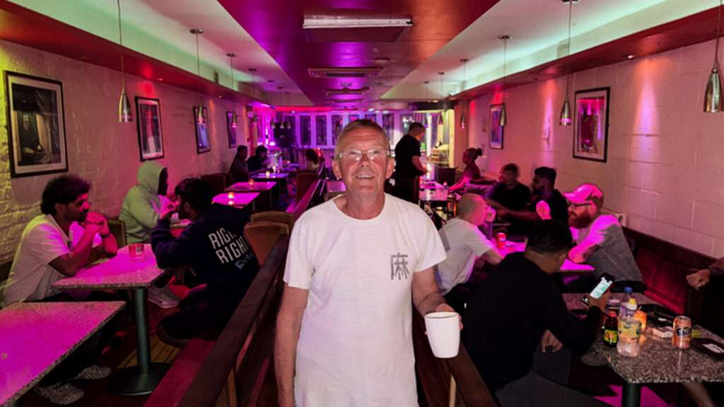 A man stands in a white t-shirt holding a tea mug with long rows of tables on either side of him. The cafe has purple lighting.