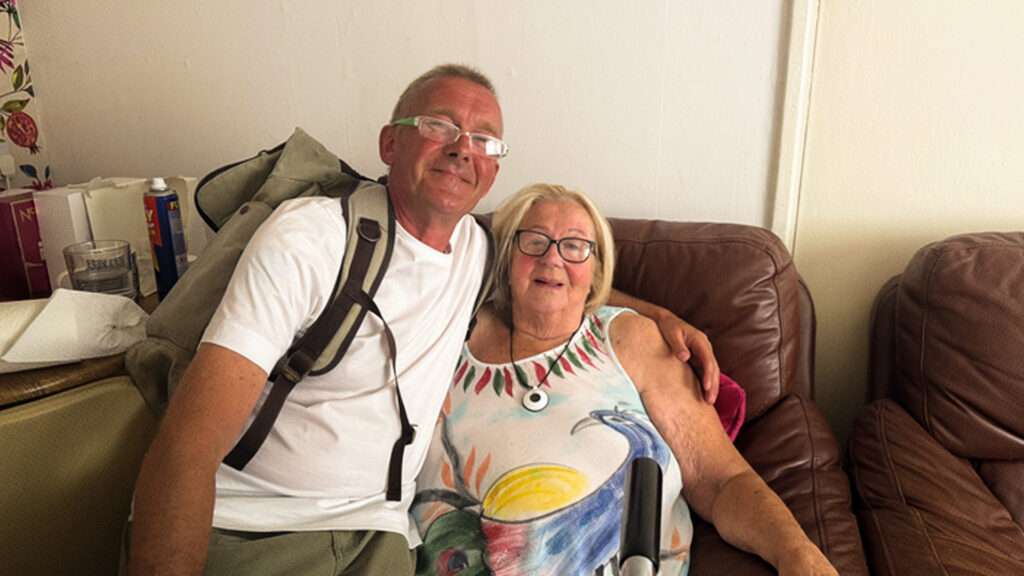 A man wearing a backpack and a white t-shirt leans on a woman sitting in a large leather chair.